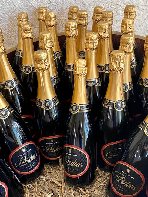 A collection of multiple bottles of sparkling wine with dark glass bodies, gold foil-wrapped necks, and black and red labels with gold accents, arranged closely together inside a cardboard box filled with wood shavings, likely prepared for home relocation or a move. The bottles are upright, and the setting appears to be indoors, possibly near an entryway or in a storage area, as part of packing and handling during a house removal service provided by Removal Van Maida Vale. The image emphasizes the careful packing of beverage items alongside other belongings in preparation for transport, illustrating the detailed process of furniture and item packing during a professional removal process.