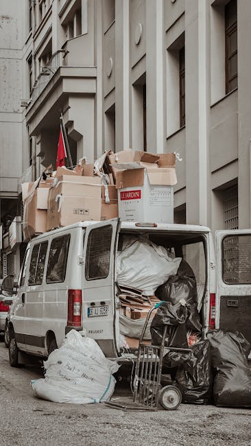 A white commercial van parked on a city street with its rear doors open, revealing various packing materials, including large plastic bags, cardboard boxes, and wrapped furniture, ready for home relocation or furniture transport. The van is situated close to a multi-storey building with a classic architectural facade, and some boxes are stacked on top of the vehicle, secured with packing tape and plastic wrap. A small hand trolley is positioned on the pavement nearby, and the scene is illuminated by natural daylight. This image illustrates the loading process involved in house removals, with a focus on efficient packing and transportation logistics, as handled by Removal Van Maida Vale.