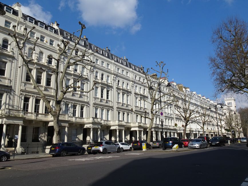 A street scene in Maida Vale showing a row of elegant, white Victorian-style terraced houses with ornate architectural details, large sash windows, and decorative balconies. Several leafless trees lin