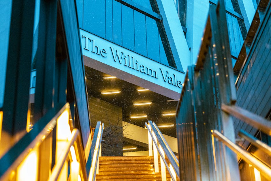 The image shows the exterior entrance of The William Vale hotel during evening hours, with the hotel name displayed prominently on a modern sign above the stairway. The steps are made of wood and lead up to glass doorways, framed by dark-colored metal railings and structures. Warm interior lighting shines through the glass, illuminating the staircase area and creating a welcoming atmosphere. The surrounding architecture features contemporary design elements, including sleek lines, dark exterior panels, and subtle lighting accents. This setting, captured in a low-angle shot, emphasizes the building's modern aesthetic and highlights the stairs leading into the hotel, which is situated in an urban environment. This visual focus on the hotel entrance aligns with themes of home or property relocation, reflecting the importance of careful planning and operational precision during house removals or furniture transport, as managed by professional removal services like Removal Van Maida Vale, especially when coordinating moving logistics.