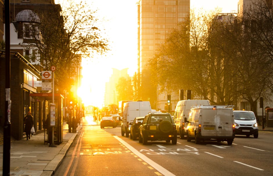 A busy urban street scene during sunset in Maida Vale, featuring a row of white and dark-colored commercial vans and smaller vehicles parked along the roadside, with a group of people waiting at a bus stop on the left. The scene includes a line of trees with autumn foliage, a bus shelter with informational signage, and a background of modern high-rise buildings illuminated by the warm glow of the setting sun. The pavement and road are visible, with marked lanes and bus stop zones, capturing the typical environment encountered during home relocation or furniture transport activities. The presence of commercial vehicles suggests ongoing loading or unloading processes, consistent with professional removals services such as those provided by Removal Van Maida Vale, situated within the context of packing, moving logistics, and traffic considerations in urban street environments.