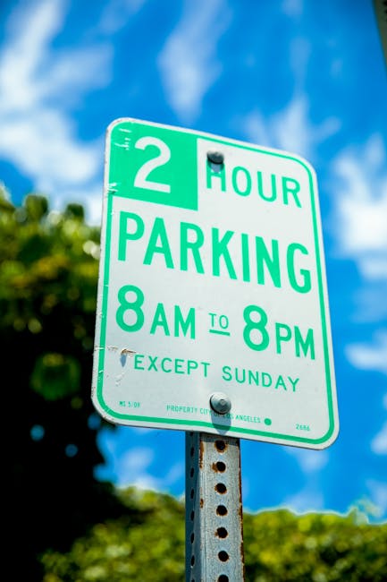 A close-up of a green parking restriction sign mounted on a metal post in an outdoor urban environment, indicating a two-hour parking limit from 8 a.m. to 8 p.m., Monday to Saturday, with no parking on Sundays. The sign is in front of a background of a partly cloudy blue sky and blurred green trees. The sign's metal post shows some signs of rust, and the sign itself has minor scratches and a small section of peeling paint. This image relates to parking regulations that may affect home relocation or furniture transport processes in Maida Vale, where Removal Van Maida Vale provides professional removals services and assistance with parking permits to facilitate efficient moving and packing operations.