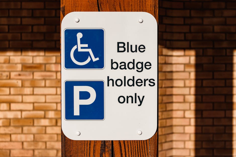 A rectangular white sign mounted on a wooden post in front of a brick wall. The top section of the sign features a blue square with a white wheelchair symbol, indicating accessibility for disabled badge holders. Below this, the sign displays black text that reads 'Blue badge holders only' next to a blue square with a white letter 'P', signifying parking restrictions exclusively for blue badge holders. The sign is part of a parking regulation setup associated with house removals or home relocation services, such as those provided by Removal Van Maida Vale. The sign appears outdoors in natural daylight, with shadows cast on the brick wall behind, and the wooden post shows visible grain and texture. This signage relates to parking rules important for removals planning in Maida Vale, ensuring accessible parking for disabled individuals during furniture transport or packing and moving activities.