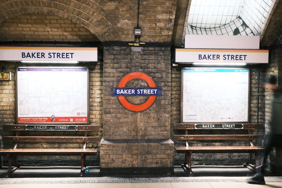 Indoor view of Baker Street underground station platform in Maida Vale, featuring two large illuminated station maps in protective frames on brick walls, with wooden benches below each map. The station name 'BAKER STREET' is displayed on signs above the maps and on a circular underground logo mounted between them. A blurred figure of a person is walking past on the right side of the image. The environment includes brick archways, a tiled ceiling, and signage related to London Underground clearly visible. The scene captures the typical setting for home relocation and furniture transport logistics, with the station environment providing context for the transportation of household items and packing materials used during removals, as managed by Removal Van Maida Vale.