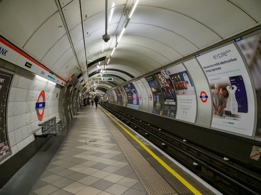 The image depicts a modern underground London Tube station, specifically Maida Vale, with the platform illuminated by overhead lighting and featuring white tiled walls and a curved ceiling. The station's iconic roundel sign with the red and blue logo is visible on the left side, alongside a black station name sign. The platform has a grey and beige diamond-patterned tiled floor with tactile paving near the edge for safety. Several passengers are standing and walking along the platform, some near the advertising panels that line the right side, displaying various posters and promotional content. The advertisements include images of electronic devices, healthcare products, and other consumer items. To the right, a section of the train tracks is visible, with the tracks running parallel to the platform. The environment appears clean and well-lit, typical of London Underground stations, and no moving or loading activities related to house removals are visible in this interior view. This image reflects the typical environment experienced during travel or within the context of planning home relocation through services like Removal Van Maida Vale.