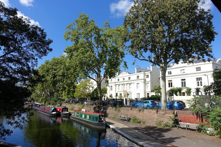 A canal with calm water reflects the surrounding trees and blue sky with a few clouds. Along the canal bank, there are several large green-leafed trees providing partial shade. On the opposite side, residential buildings painted in white are visible, with parked cars including blue and silver vehicles lining the street. A brick wall separates the sidewalk from the canal, with several wooden benches positioned along the walkway. In the foreground, small boats with dark hulls and red, green, and white details are moored at the canal edge, with some boats appearing to be used for sightseeing or leisure. The scene captures a peaceful urban environment typical of a residential area in Maida Vale, suitable for house removals, furniture transport, or packing and moving activities by companies such as Removal Van Maida Vale, which may handle relocation services in the vicinity.