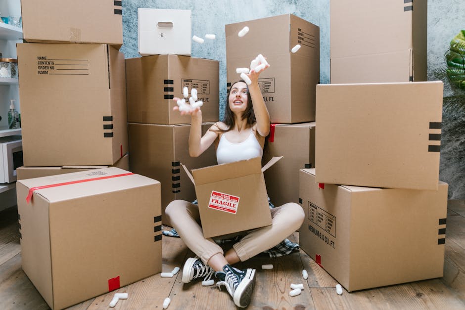 A woman sitting cross-legged on a wooden floor inside a room, surrounded by stacked cardboard boxes of various sizes, some sealed with red tape and labeled with contents and fragile symbols. She is smiling and tossing packing peanuts into the air while emerging from a partially open box that is lined with a 'Fragile' sticker. The room has a light blue textured wall, and in the background, there is a small side table with a bottle and potted plant. In the context of house removals and packing and moving processes, the scene depicts a home relocation scenario with furniture and contents prepared for transport by Removal Van Maida Vale, highlighting the packing stage of a house move.