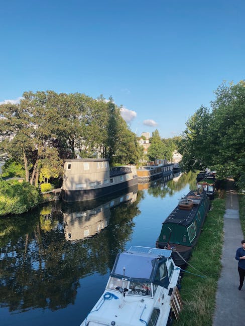 A canal with calm water reflects the surrounding trees and blue sky with a few clouds. Along the canal bank, there are several large green-leafed trees providing partial shade. On the opposite side, residential buildings painted in white are visible, with parked cars including blue and silver vehicles lining the street. A brick wall separates the sidewalk from the canal, with several wooden benches positioned along the walkway. In the foreground, small boats with dark hulls and red, green, and white details are moored at the canal edge, with some boats appearing to be used for sightseeing or leisure. The scene captures a peaceful urban environment typical of a residential area in Maida Vale, suitable for house removals, furniture transport, or packing and moving activities by companies such as Removal Van Maida Vale, which may handle relocation services in the vicinity.