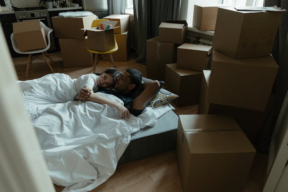 A spacious, well-lit bedroom with light-colored walls and a beige carpet. The bed, positioned against the left wall, features a tufted beige headboard and is made with white bedding. To the left of th