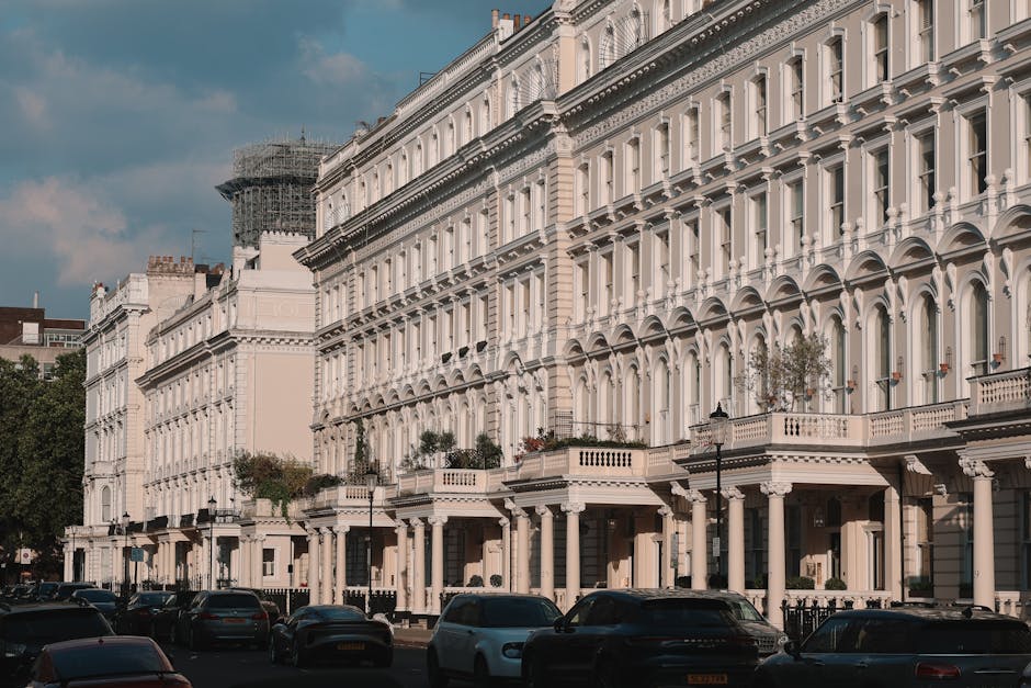 A street scene outside a row of white, Victorian-style terraced houses with ornate facades, large windows, and decorative molding, typical of the Maida Vale area. Several cars are parked along the pavement, and the road is illuminated by classic-style street lamps. The buildings feature small front gardens with potted plants and iron railings, and some windows have window boxes. The image captures clear, daylight conditions with a partly cloudy sky overhead. In the foreground, part of a loading area adjacent to the properties shows a black van used for home relocation services, with visible cardboard boxes, plastic wrapping, and furniture including chairs and small tables being loaded or prepared for transport. The scene illustrates the process of furniture transport and packing involved in house removals, as provided by Removal Van Maida Vale, emphasizing the logistics of loading household items onto the van for a professional move in the Maida Vale district.