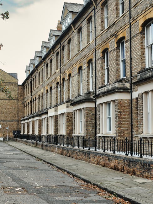 A street scene outside a row of white, Victorian-style terraced houses with ornate facades, large windows, and decorative molding, typical of the Maida Vale area. Several cars are parked along the pavement, and the road is illuminated by classic-style street lamps. The buildings feature small front gardens with potted plants and iron railings, and some windows have window boxes. The image captures clear, daylight conditions with a partly cloudy sky overhead. In the foreground, part of a loading area adjacent to the properties shows a black van used for home relocation services, with visible cardboard boxes, plastic wrapping, and furniture including chairs and small tables being loaded or prepared for transport. The scene illustrates the process of furniture transport and packing involved in house removals, as provided by Removal Van Maida Vale, emphasizing the logistics of loading household items onto the van for a professional move in the Maida Vale district.