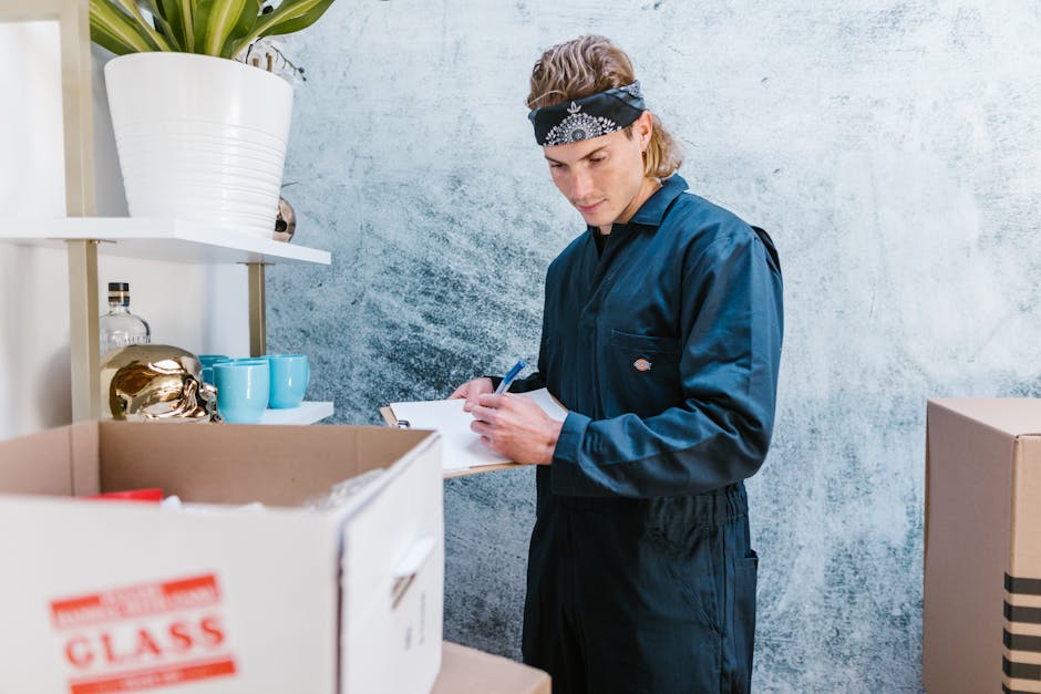 A professional removalist wearing a dark blue uniform and a black bandana with white patterns is inside a room, engaged in a home relocation process. He is standing next to a cardboard box filled with packing materials, with some boxes on a nearby shelf and others on a table or surface. The man is holding a clipboard and pen, likely taking inventory or notes, indicative of organized packing and moving procedures. The room features a textured, light-colored wall with shelves that hold decorative items such as a white ceramic pot with green plants, blue cups, and a metallic decorative object. The environment is well-lit, allowing clear visibility of the objects and the removalist’s actions, emphasizing the packing and loading aspects of furniture transport and house removals, as conducted by Removal Van Maida Vale in the context of urgent same-day moves.