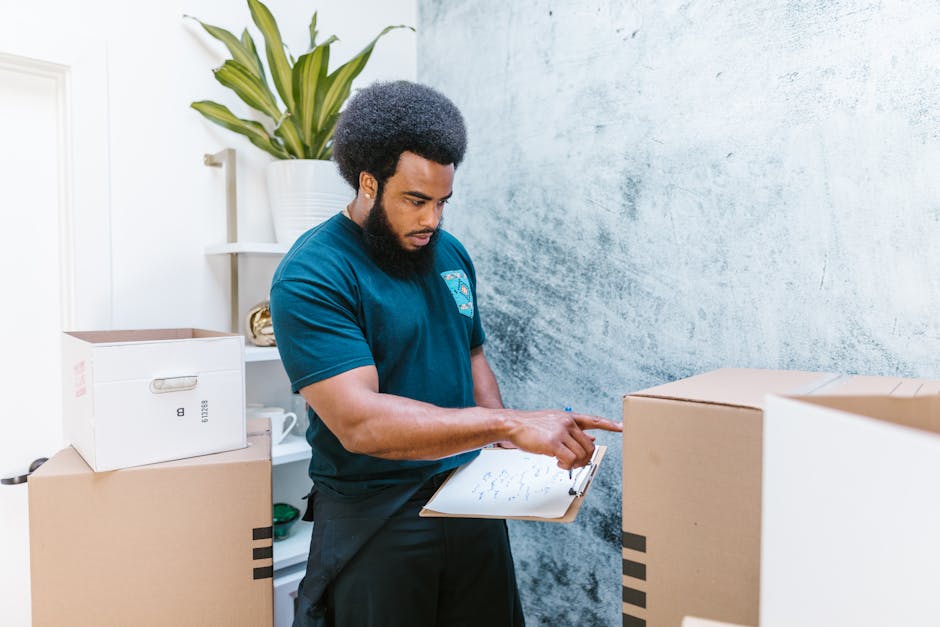 A person wearing a yellow t-shirt and blue jeans is standing inside a room filled with packed cardboard boxes of various sizes, some open and some closed, ready for home relocation. They are holding a clipboard and a black marker, appearing to take notes or check off items in a moving checklist. The room has a light-colored wall and wooden flooring, with a large green potted plant placed in a black pot on the left side. Several cardboard boxes are stacked behind the individual, suggesting an ongoing packing or loading process as part of furniture transport. The scene is well-lit, highlighting the practical steps involved in packing and moving, which are integral to professional removals services like those offered by Removal Van Maida Vale.