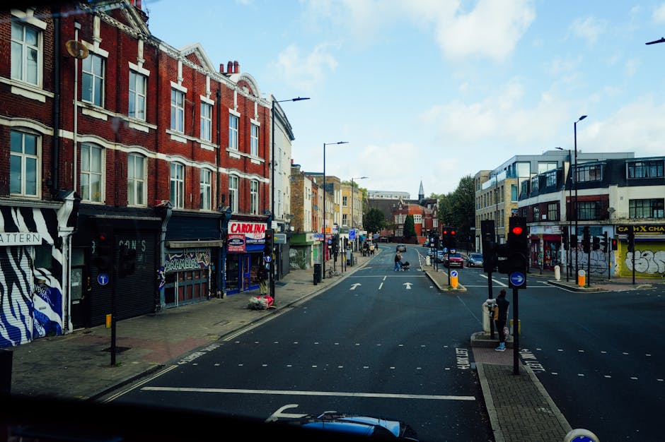 A view of Sutherland Avenue in Maida Vale showing a busy street with commercial and residential buildings on both sides. To the left, there are shops with large windows and signage, some with black and white striped awnings and graffiti-style artwork on the shop fronts. To the right, modern apartment buildings with glass balconies and some Graffiti art on the lower walls. The street is occupied by a few pedestrians waiting at traffic lights, and a vehicle's dashboard is visible at the bottom of the image, indicating the photo was taken from inside a vehicle. The road has clear white lane markings, traffic lights, and bollards at pedestrian crossings, with cars visible in the distance. This setting captures a typical urban scene suitable for a home relocation or furniture transport process by Removal Van Maida Vale, with the area prepared for loading and unloading of household goods, including possible use of trolleys or other equipment for moving furniture and packed boxes, situated near the pavement and shop entrances.