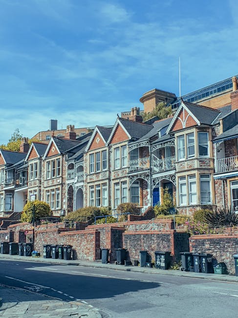 A row of historic terraced houses with bay windows, decorative ironwork, and brick facades situated on Sutherland Avenue in Maida Vale. The houses are built on an elevated level with small front gardens enclosed by low brick walls, some with shrubbery and potted plants. A parked removal van from Removal Van Maida Vale is adjacent to the curb, with the sliding side door open, revealing a loading area with cardboard boxes, wrapped furniture, and plastic covers being prepared for a home relocation. The van is positioned close to the property entrance, which features a pathway leading to the front door, and the scene is lit by bright daylight under a clear blue sky. The environment includes a paved street with black wheelie bins lined up along the pavement, and the overall setting illustrates the logistics involved in furniture transport and packing for house removals in the Maida Vale area.
