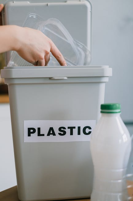 A person dressed in orange work overalls is standing on a gray indoor floor, holding two large blue plastic bags filled with waste or recyclables, with the bags hanging down by their sides. The indivi