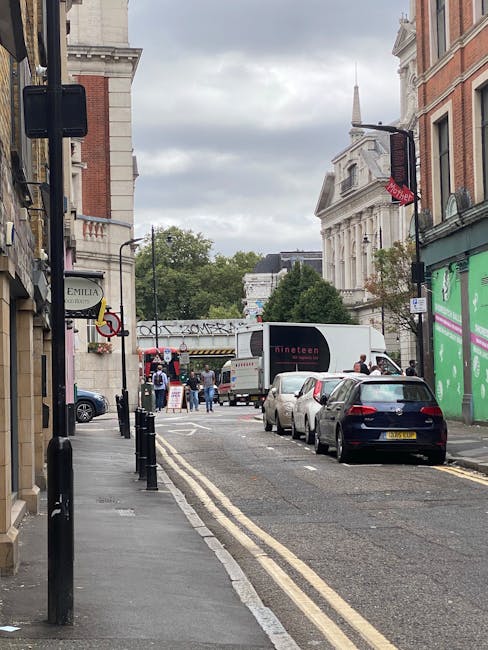 A street scene in Maida Vale showing a narrow road with parked cars along the right side, including a black hatchback closest to the camera, and a white van slightly further down. On the left side, there is a sidewalk with black bollards separating it from the road, and a lamppost with signage attached. In the distance, a large white moving truck with the branding 'Dineet' is positioned on the street, with people nearby possibly involved in home relocation activities. The buildings along the street are a mix of brick and stone architecture, with some featuring large windows and decorative details. Overhead, the sky is overcast with grey clouds, and trees are visible further along the street, providing greenery. The scene depicts the logistics involved in furniture transport and packing during moving services provided by Removal Van Maida Vale, with signs of a busy residential area prepared for a house move, adhering to council regulations for loading and unloading.