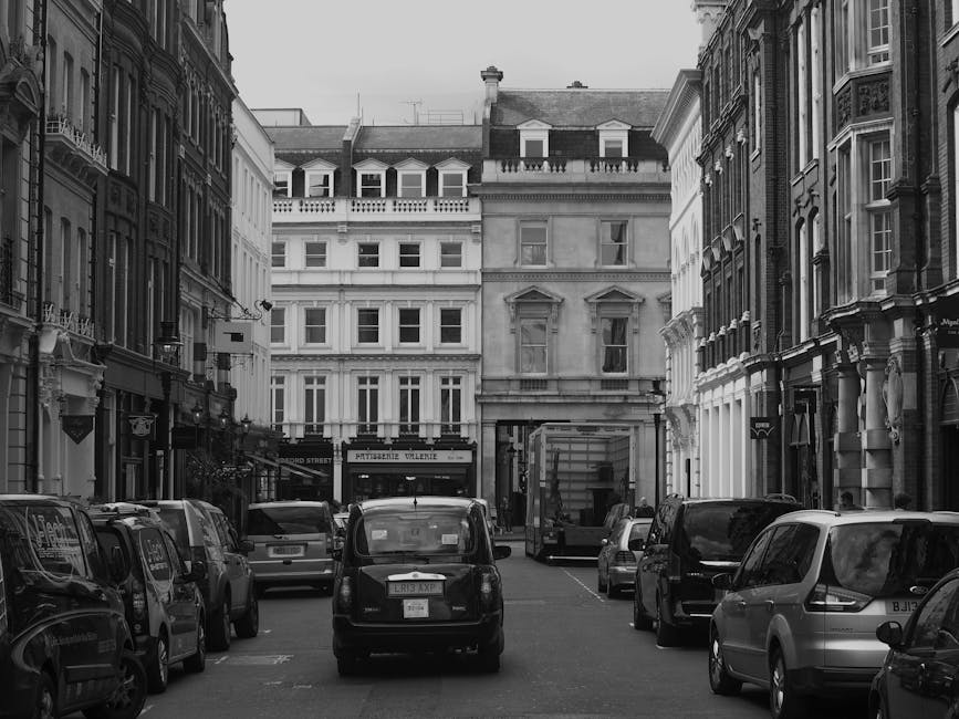 A street scene in Maida Vale showing a narrow road with parked cars along the right side, including a black hatchback closest to the camera, and a white van slightly further down. On the left side, there is a sidewalk with black bollards separating it from the road, and a lamppost with signage attached. In the distance, a large white moving truck with the branding 'Dineet' is positioned on the street, with people nearby possibly involved in home relocation activities. The buildings along the street are a mix of brick and stone architecture, with some featuring large windows and decorative details. Overhead, the sky is overcast with grey clouds, and trees are visible further along the street, providing greenery. The scene depicts the logistics involved in furniture transport and packing during moving services provided by Removal Van Maida Vale, with signs of a busy residential area prepared for a house move, adhering to council regulations for loading and unloading.